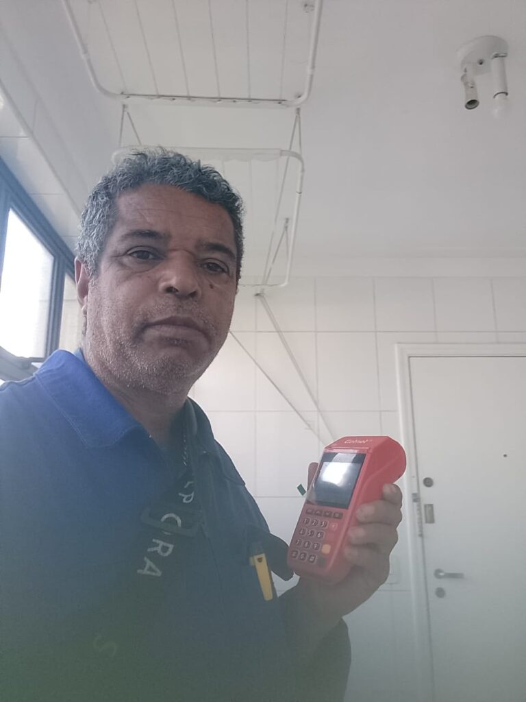 Seu Domingos Eletricista vestido com macacão azul segurando a maquininha vermelhinha GetNet, pronto para receber pagamentos de serviços elétricos.