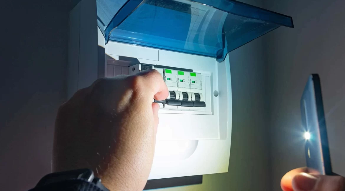 Mão mexendo no quadro de luz com lanterna de celular durante queda de energia em um cômodo escuro