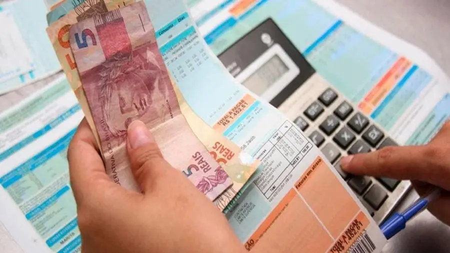 Pessoa analisando fatura da conta de energia elétrica com notas e moedas de real brasileiro